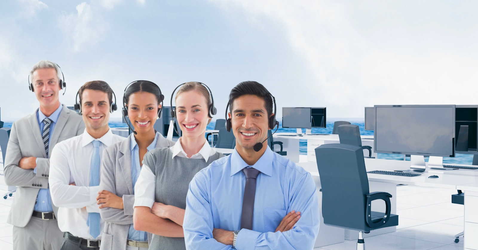 Cinco agentes de servicio al cliente sonrientes con auriculares, de pie frente a un escritorio con monitores de computadora.