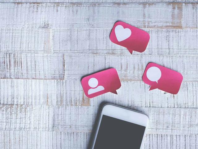 White smartphone with dark screen, three pink speech bubbles with white icons for like, comment, and profile on wood.