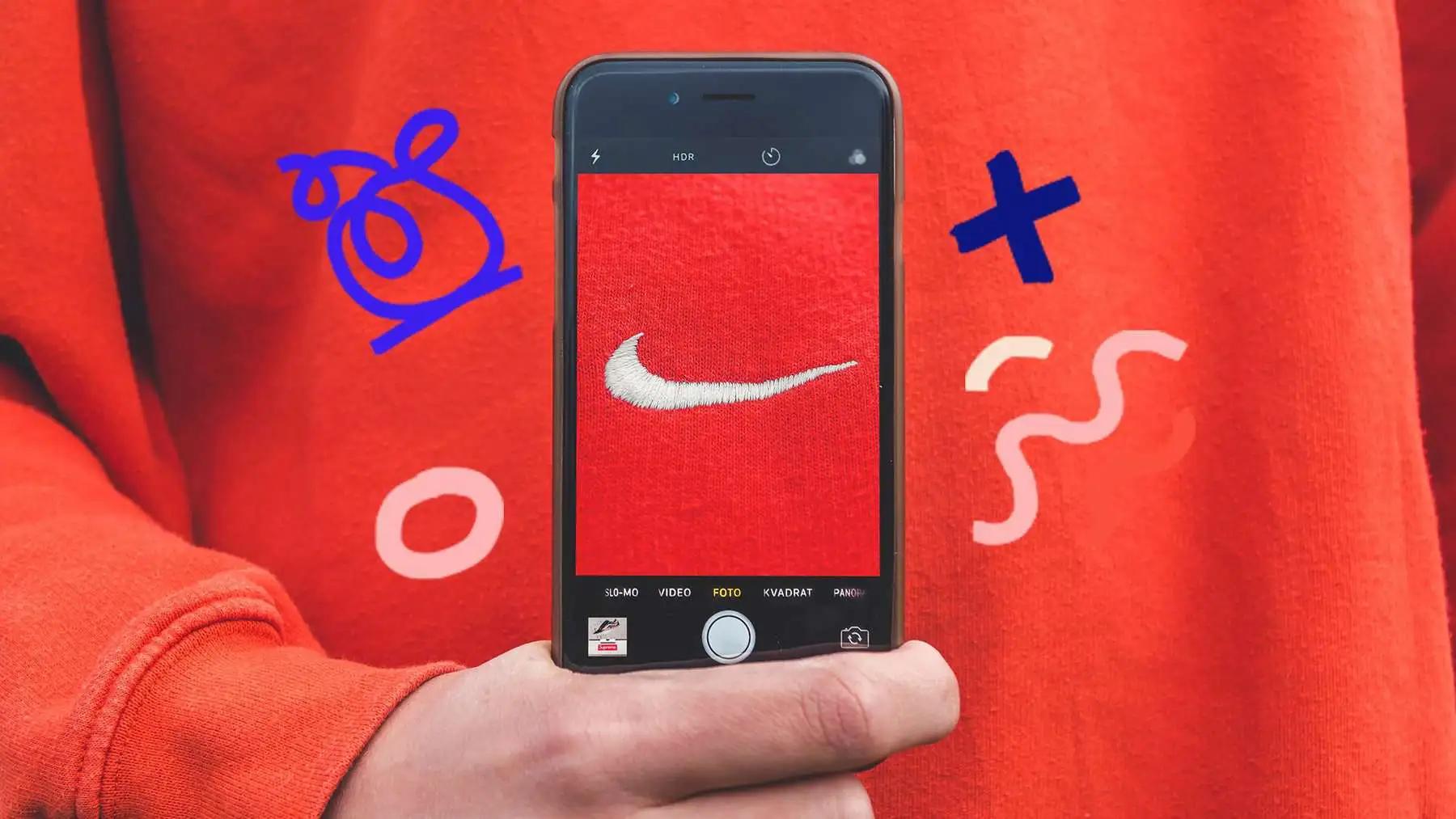 A hand holds a phone photographing a Nike swoosh on an orange sweatshirt, with doodle graphics around it