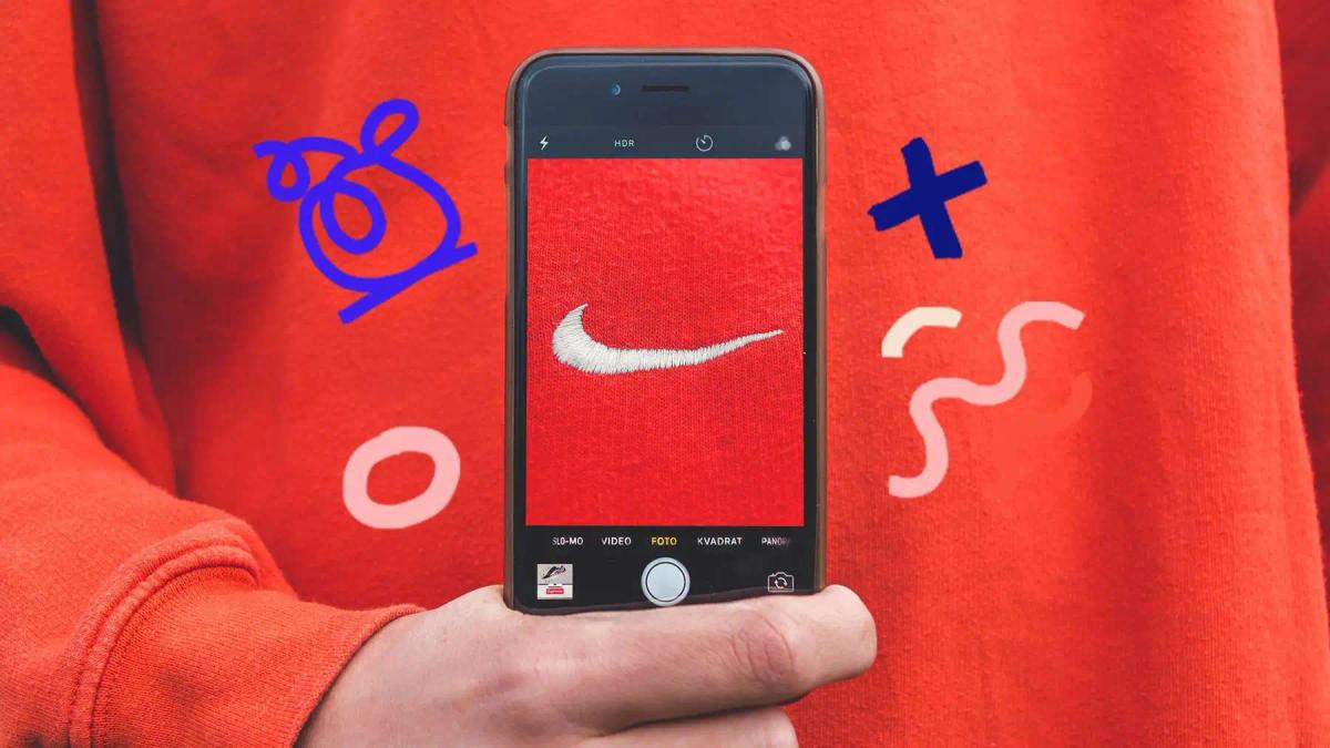 Una mano sostiene un telefono fotografiando el logo de Nike en una sudadera naranja, con garabatos alrededor
