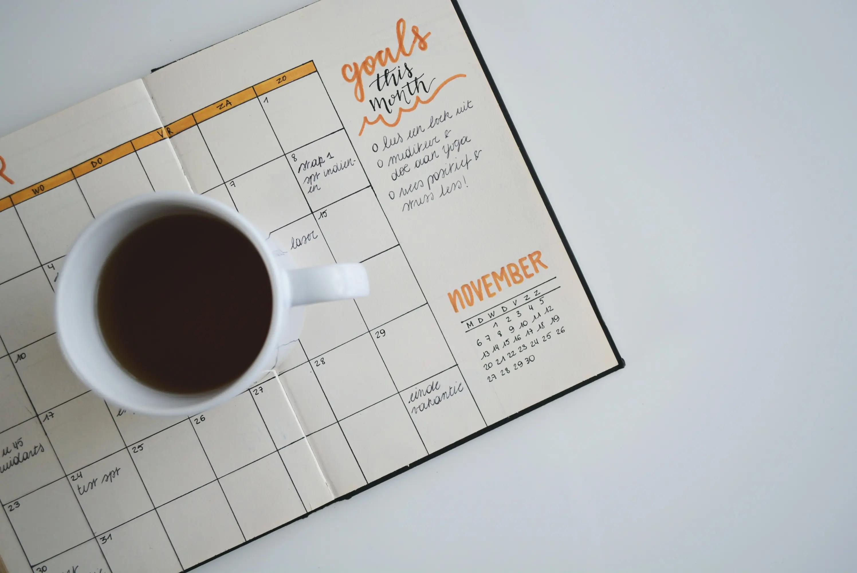 A cup of coffee sits on an open paper planner with a monthly calendar grid and handwritten goals