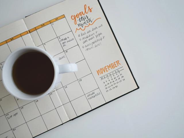 A cup of coffee sits on an open paper planner with a monthly calendar grid and handwritten goals