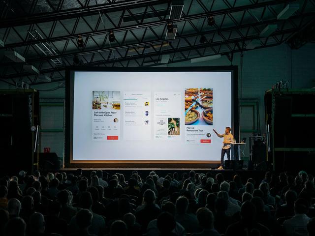 A presenter on stage gestures at a large screen showing app interface slides to a seated audience