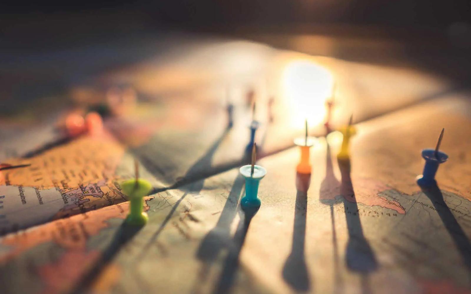 Colorful pushpins stuck in a paper map, casting long shadows in warm light