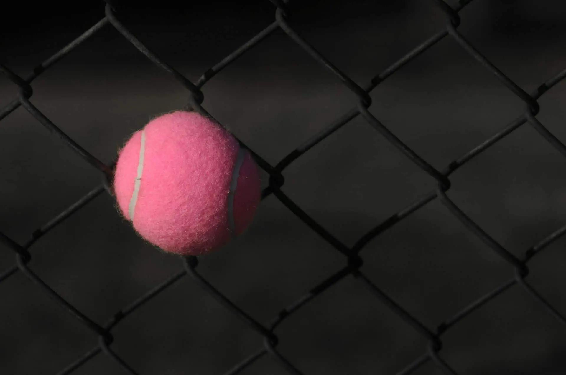 A pink tennis ball wedged in the diamond gaps of a chain-link fence