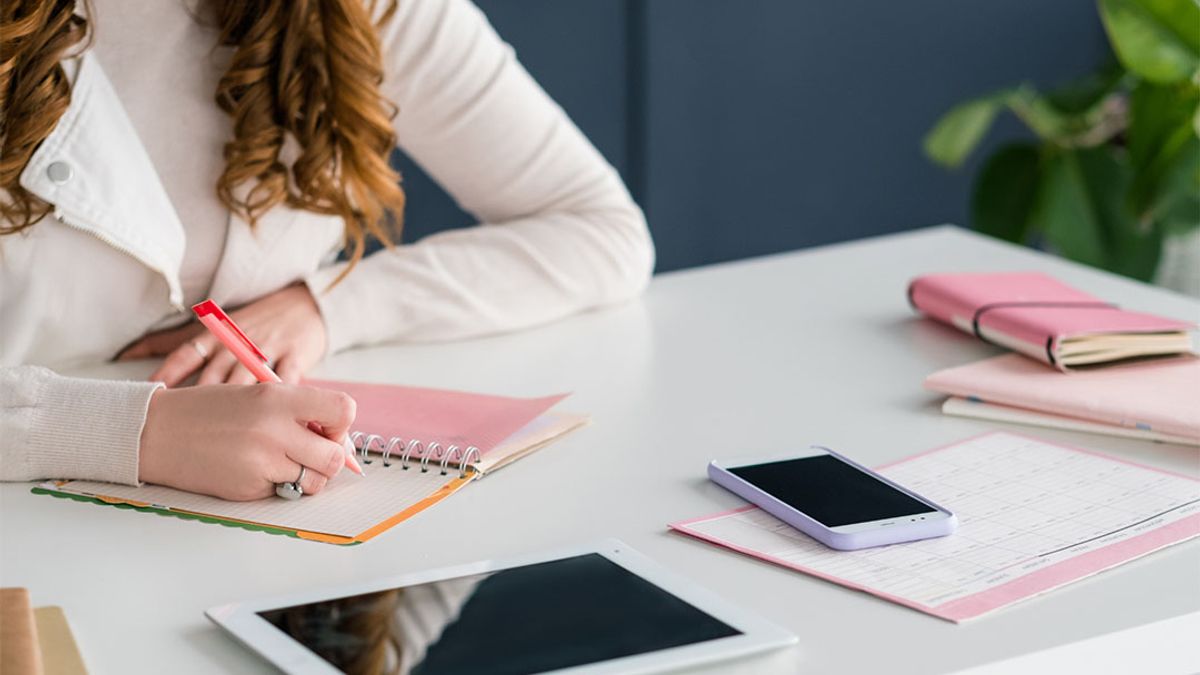 Person with long brown hair writing in a spiral notebook with a red pen. A tablet, phone, and pink notebooks are on the white desk.