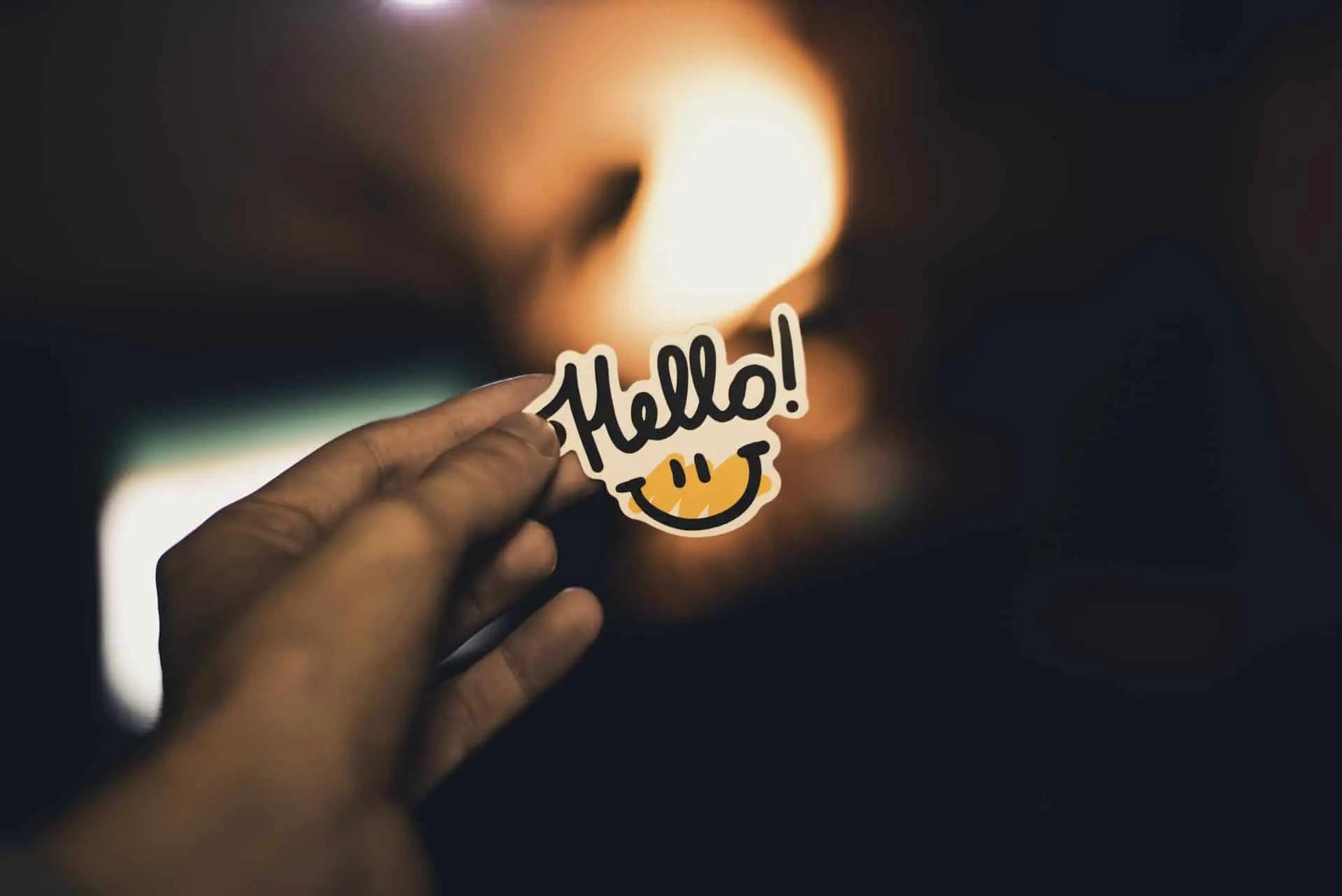 A hand holds a sticker reading Hello with a smiley face, against a dim, warmly lit background