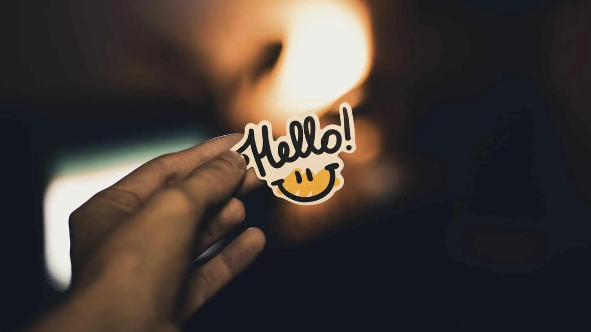 A hand holds a sticker reading Hello with a smiley face, against a dim, warmly lit background
