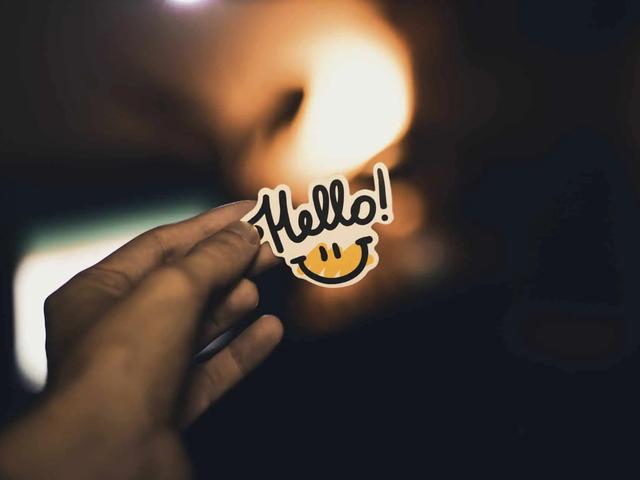A hand holds a sticker reading Hello with a smiley face, against a dim, warmly lit background