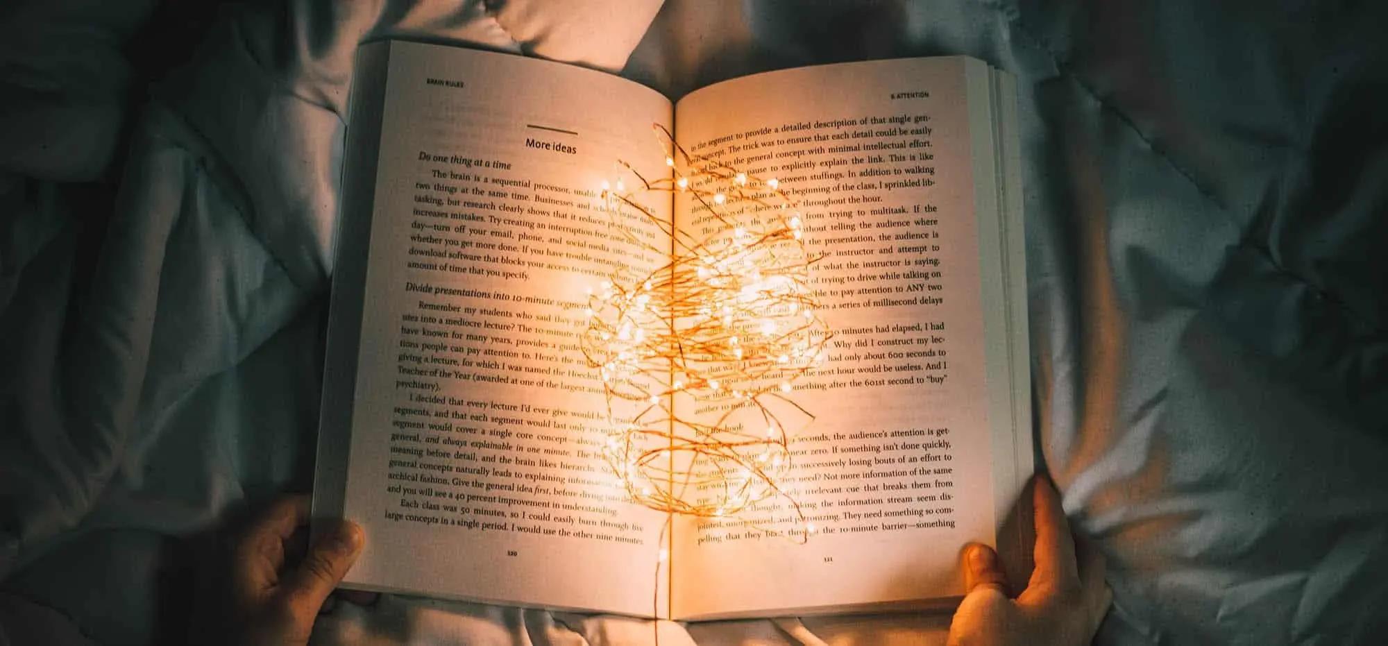 Hands holding an open book in bed with glowing fairy lights draped across the pages
