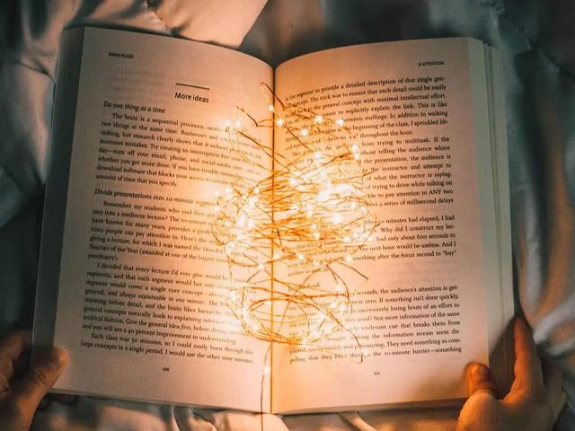 Unas manos que sostienen un libro abierto en la cama, con unas lucecitas de colores que se deslizan por las páginas