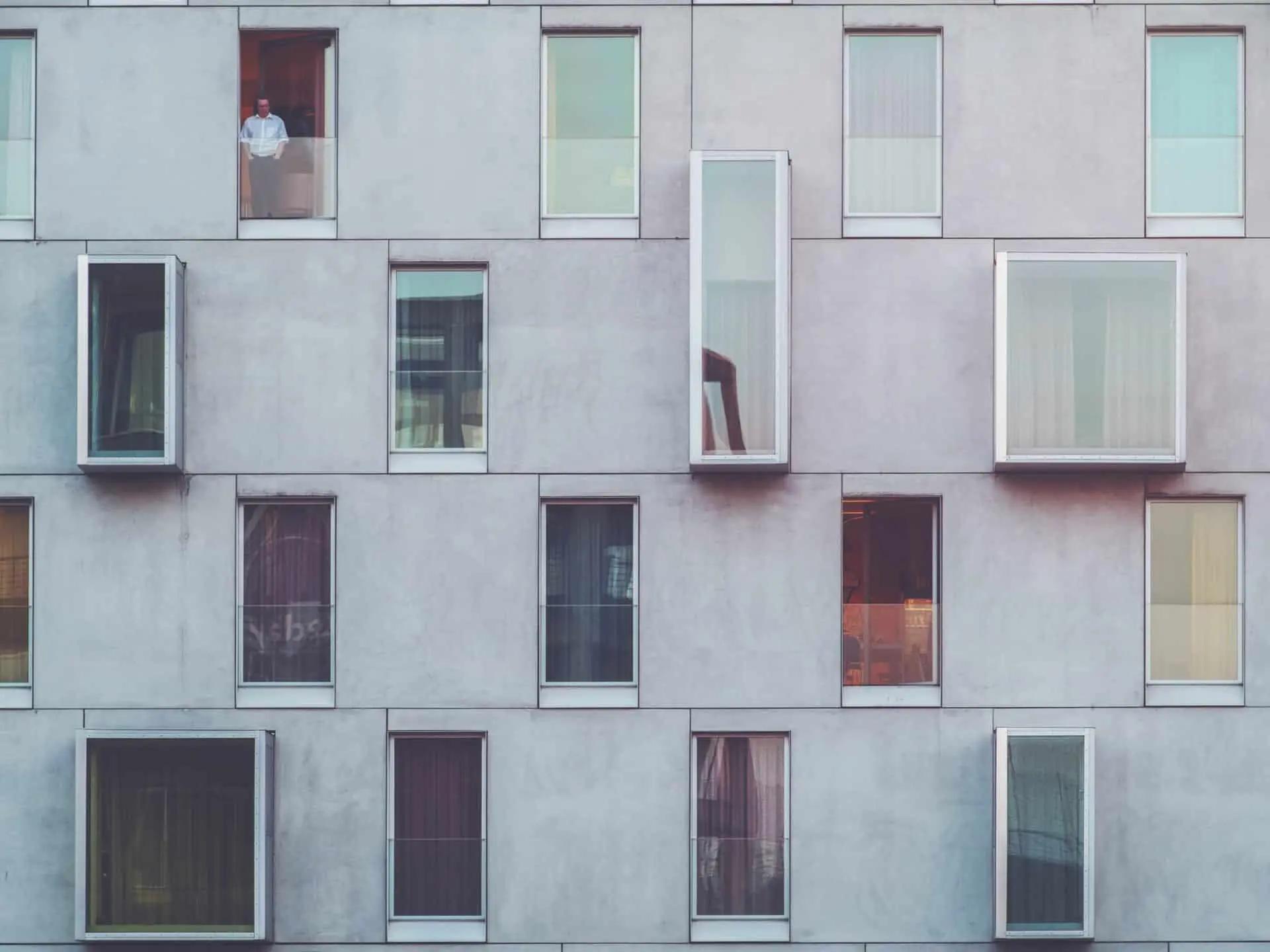 Gray building facade with many windows of different shapes and sizes. A man in a white shirt stands in one window.