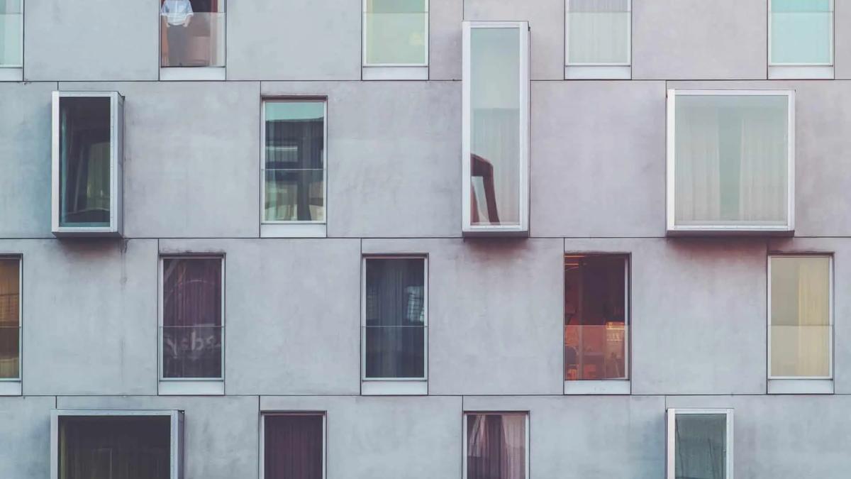 Gray building facade with many windows of different shapes and sizes. A man in a white shirt stands in one window.