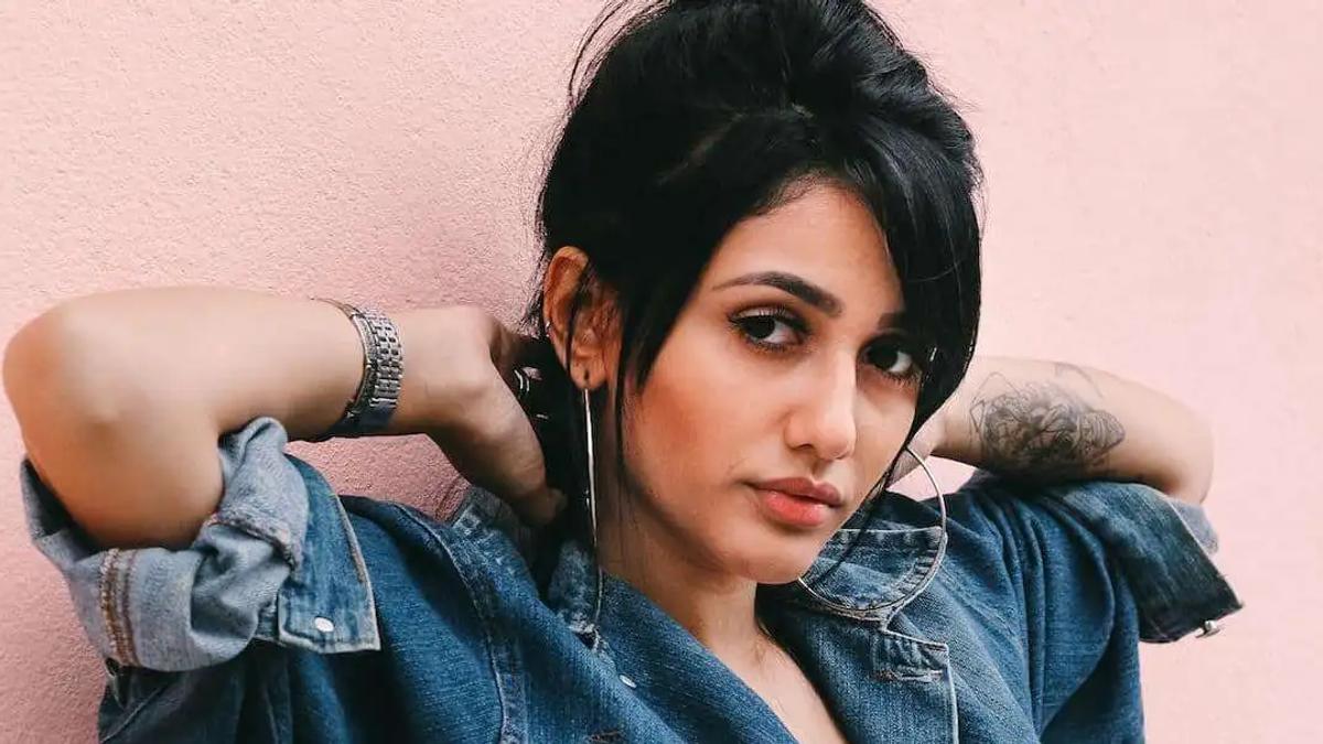 Woman in a denim jacket and hoop earrings posing against a pink wall