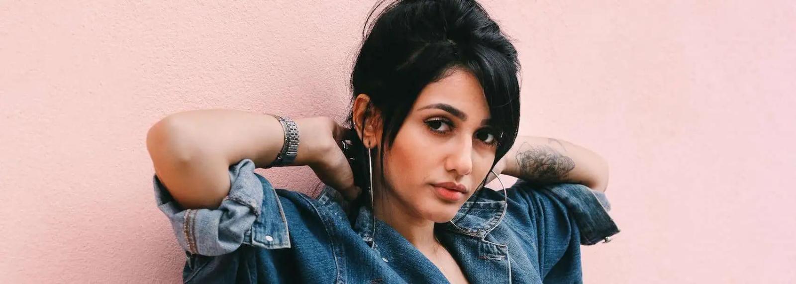 Woman in a denim jacket and hoop earrings posing against a pink wall