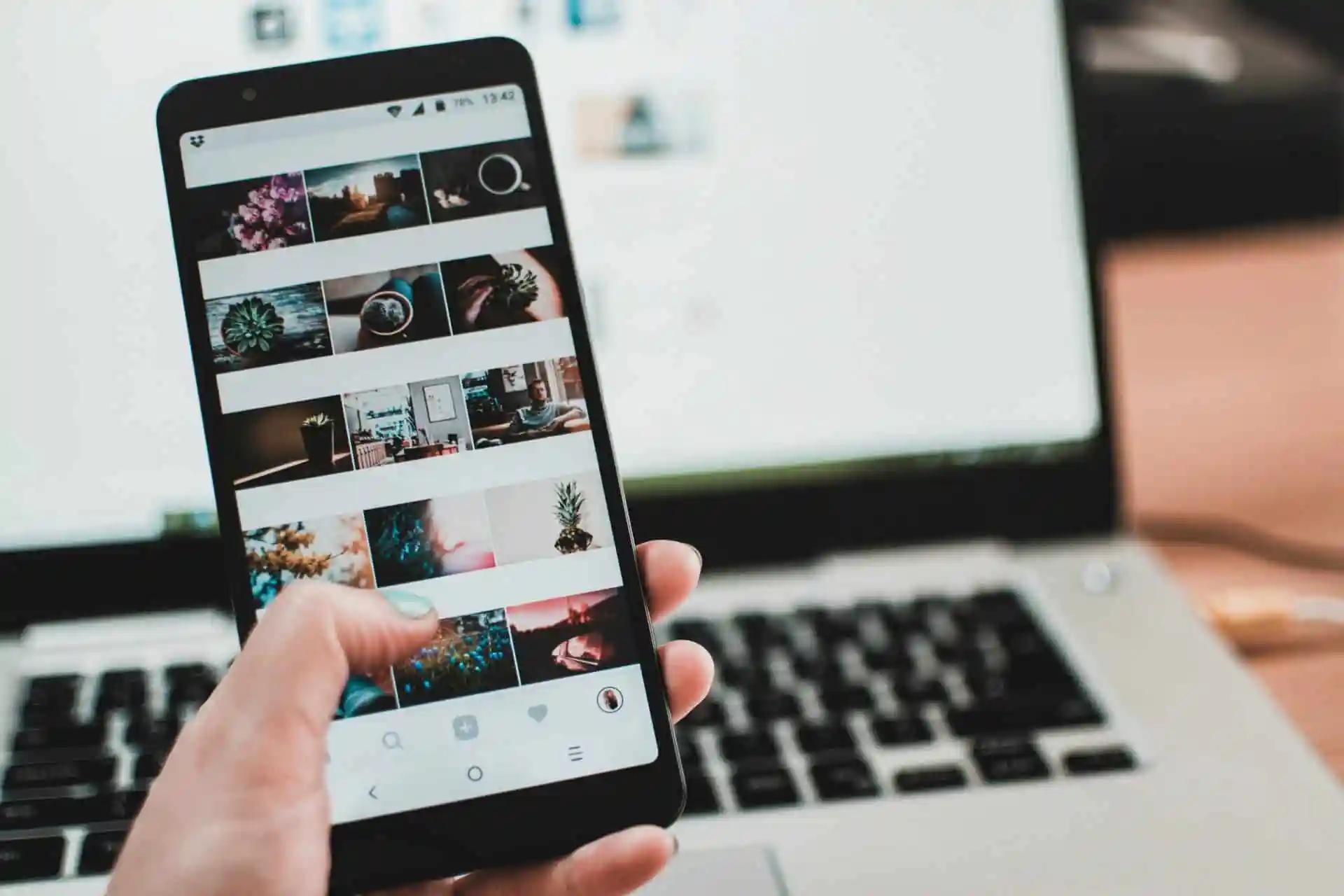 A hand holds a phone showing an Instagram grid of photos, with a laptop keyboard in the background