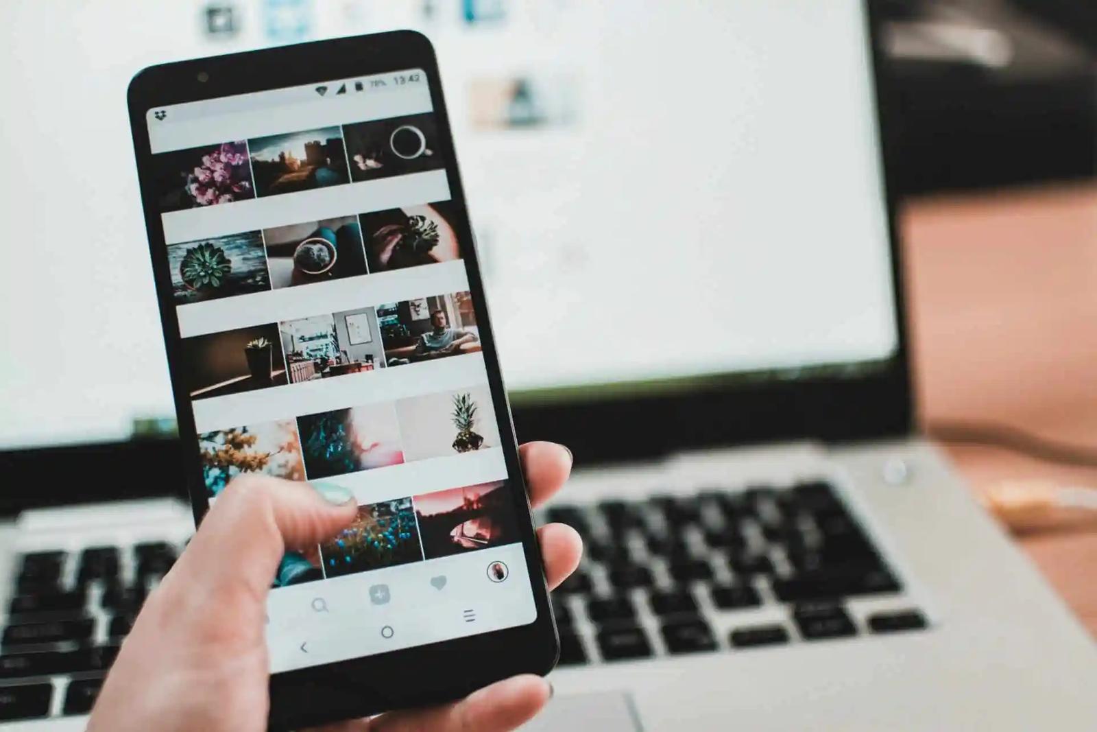 Una mano sostiene un telefono con una cuadricula de fotos de Instagram, con un teclado de laptop de fondo