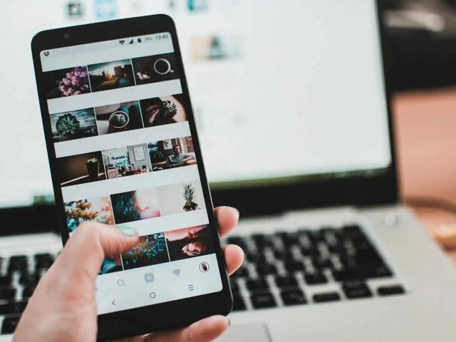 Una mano sostiene un telefono con una cuadricula de fotos de Instagram, con un teclado de laptop de fondo