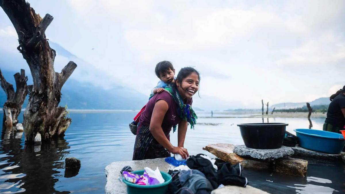 Mujer sonriente con un niño a la espalda, lavando ropa en un lago con montañas y árboles muertos al fondo