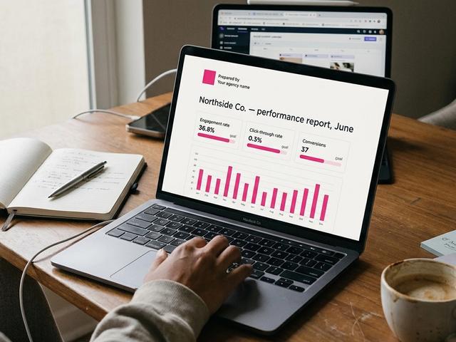 Laptop on a real desk showing a clean one-page client social media report with KPI tiles and a pink bar chart.