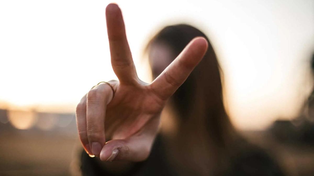 A woman holds up a peace sign toward the camera against a sunset backdrop