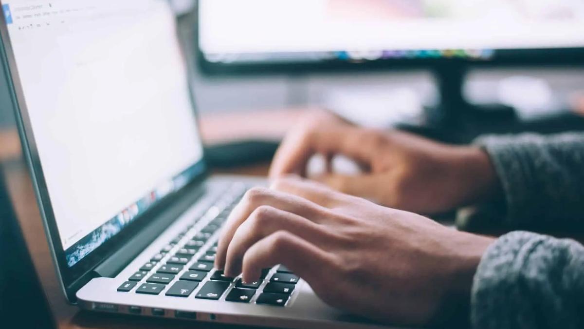 Hands typing on a silver laptop keyboard, with a blurred monitor in the background. Warm, focused lighting.