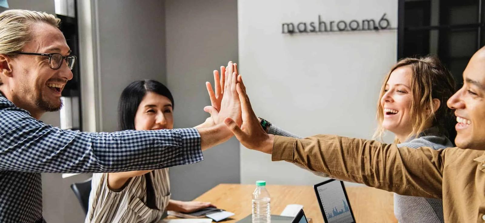 Four coworkers high-fiving across an office table with laptops and a bottle of water