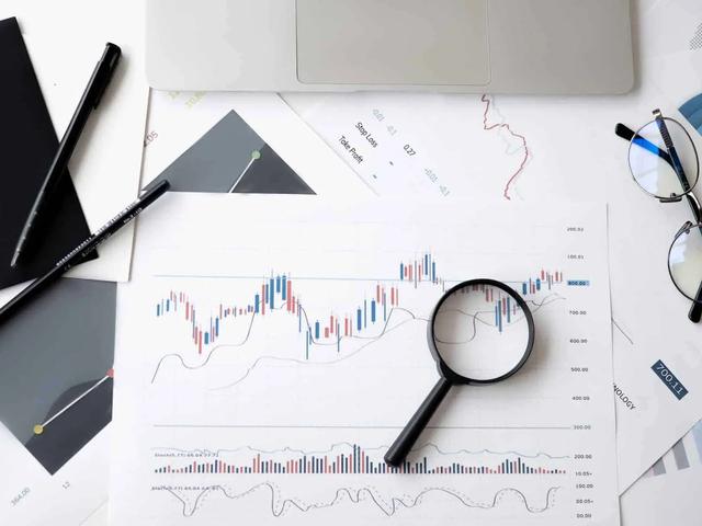 Overhead view of a laptop, papers with charts and graphs, a magnifying glass, pens, and glasses on a white desk