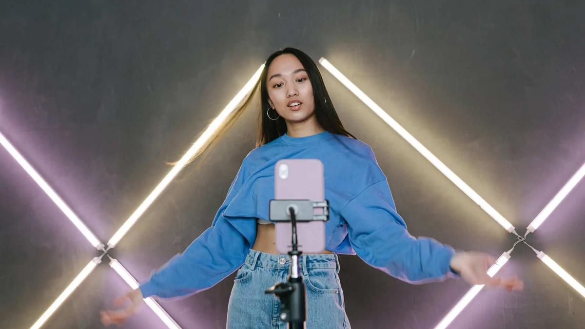 Young woman in blue top and jeans recording a video with a smartphone on a tripod, neon lights in background