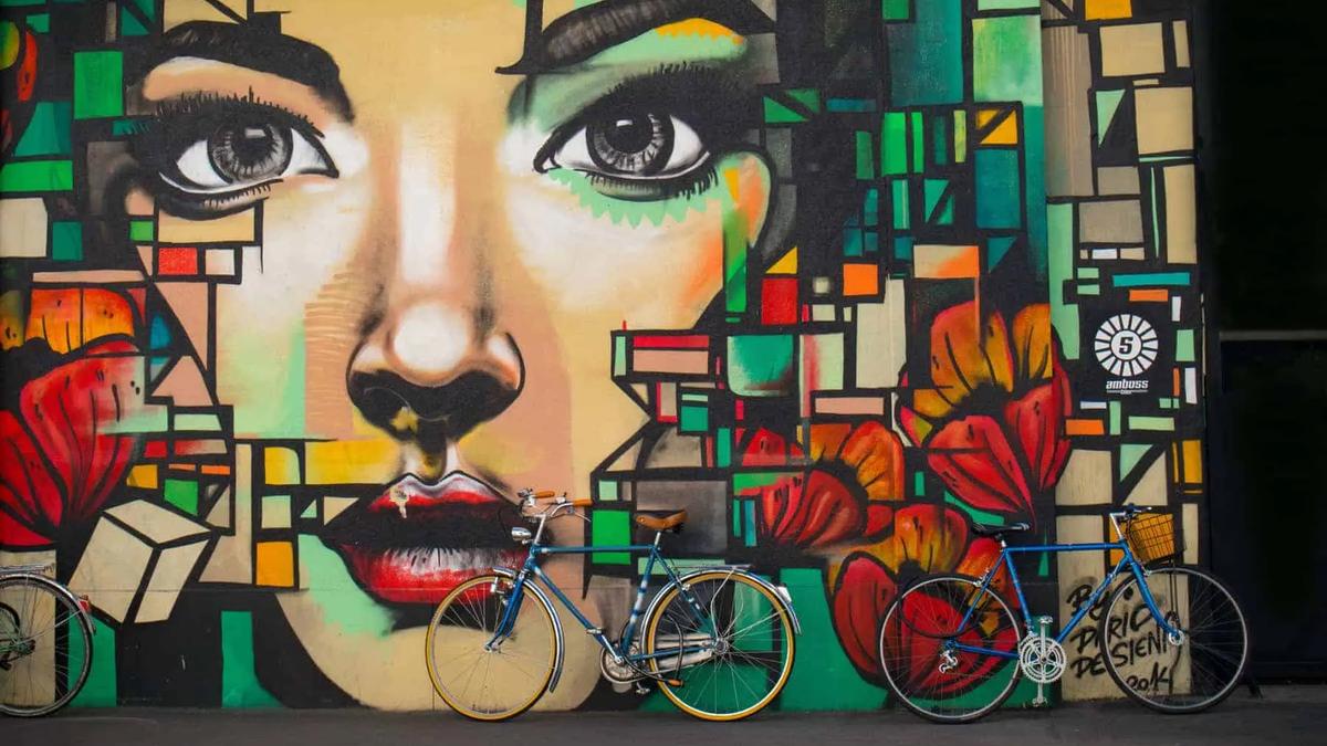 Mural colorido del rostro de una mujer con formas abstractas y flores rojas. Tres bicicletas apoyadas en la pared.