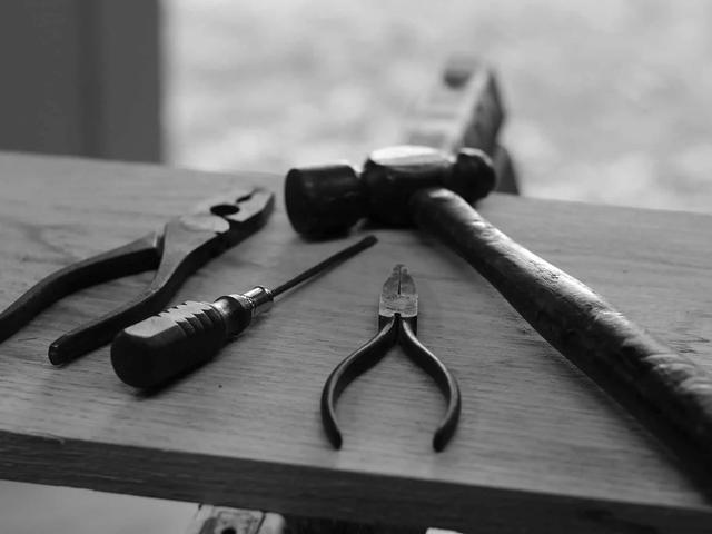 Black and white photo of pliers, a screwdriver and a hammer laid on a wooden workbench
