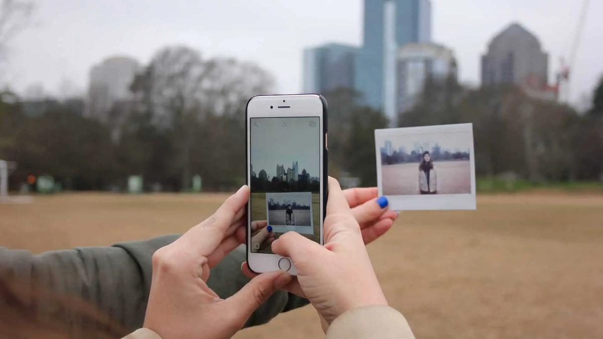 Two hands holding an iPhone and a small instant photo, with a city skyline and trees in the background on a cloudy day