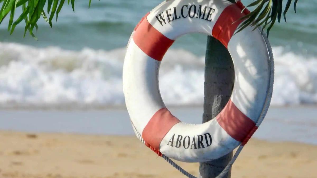 A red and white life ring reading 'Welcome Aboard' hangs on a post near a beach.