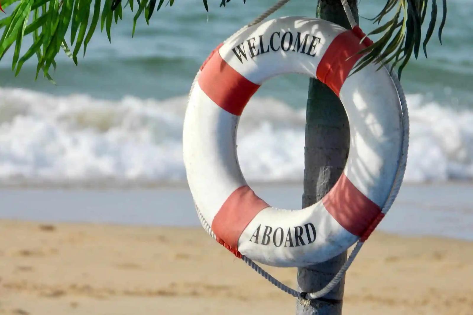 Un salvavidas rojo y blanco con la frase 'Welcome Aboard' colgado junto a una playa.