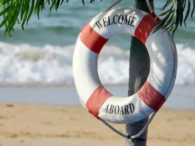 A red and white life ring reading 'Welcome Aboard' hangs on a post near a beach.