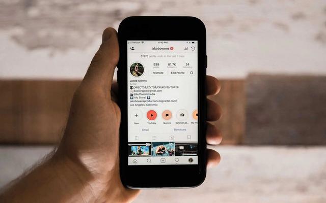 Hand holding a black smartphone displaying an Instagram profile page for Jakob Owens, white background, neutral mood.