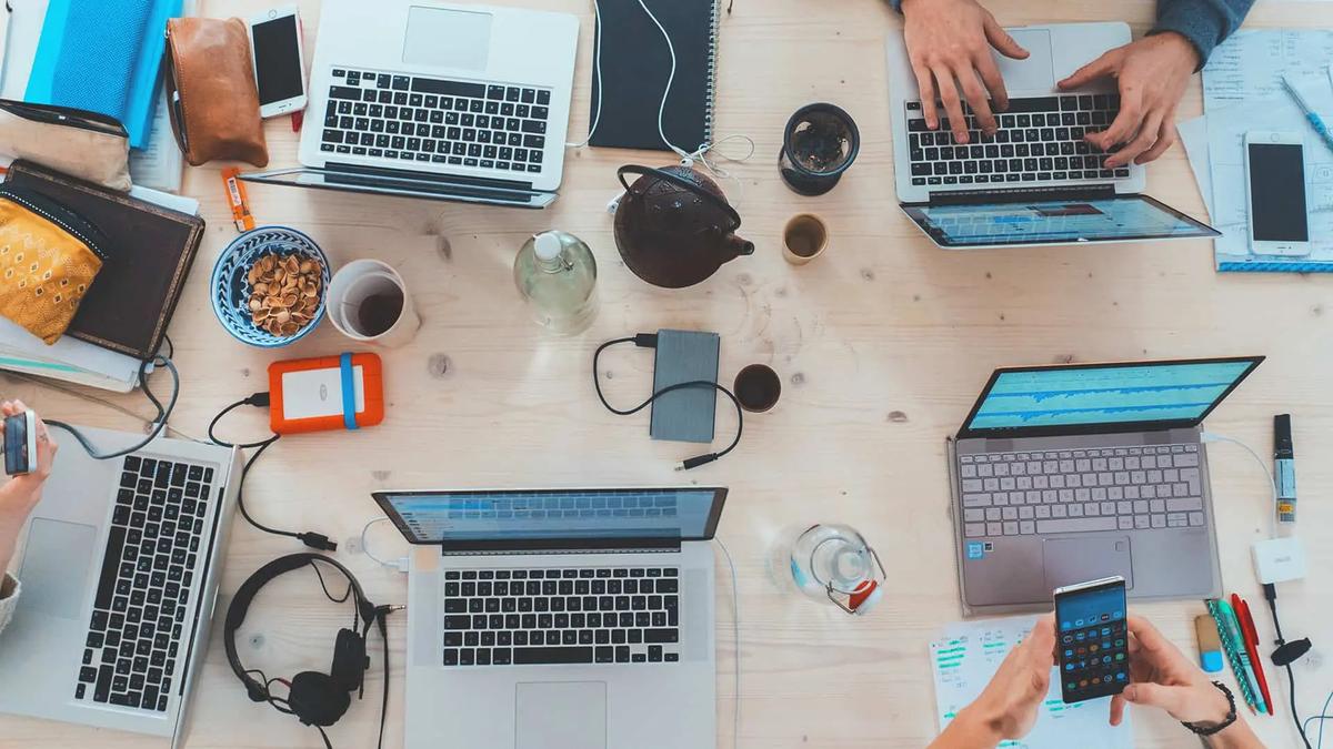 Overhead view of a wooden desk with several laptops, phones, and hands typing among cups and cables