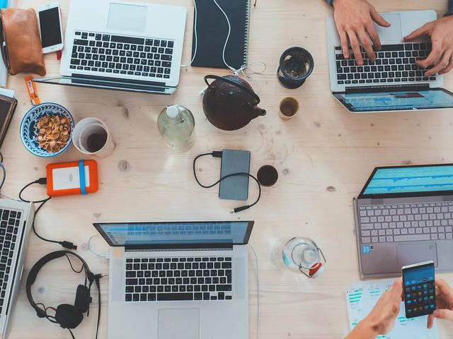 Overhead view of a wooden desk with several laptops, phones, and hands typing among cups and cables