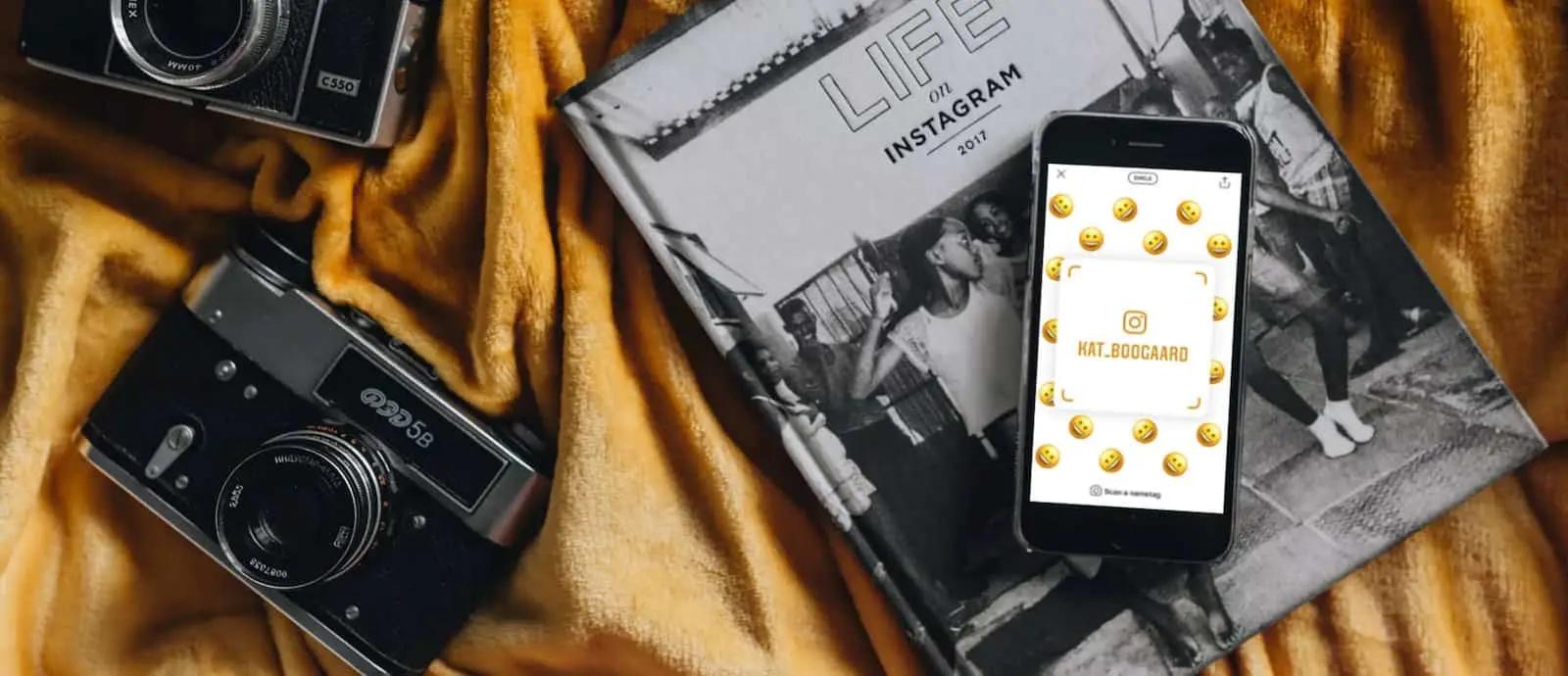 Un teléfono con una tarjeta de identificación de Instagram rodeada de emojis, sobre un libro junto a dos cámaras