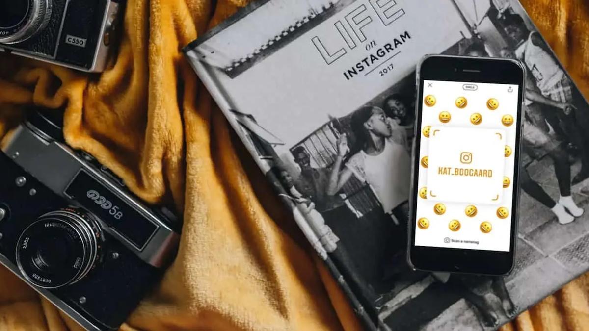 A phone showing an Instagram nametag surrounded by emoji, resting on a book beside two cameras