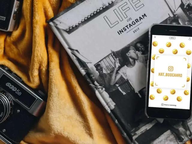 A phone showing an Instagram nametag surrounded by emoji, resting on a book beside two cameras