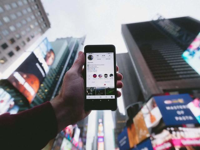Hand holding a smartphone displaying an Instagram profile, surrounded by tall buildings and bright billboards