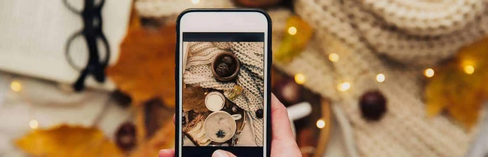 Una mano sostiene un telefono fotografiando una escena otonal con cafe y pinas