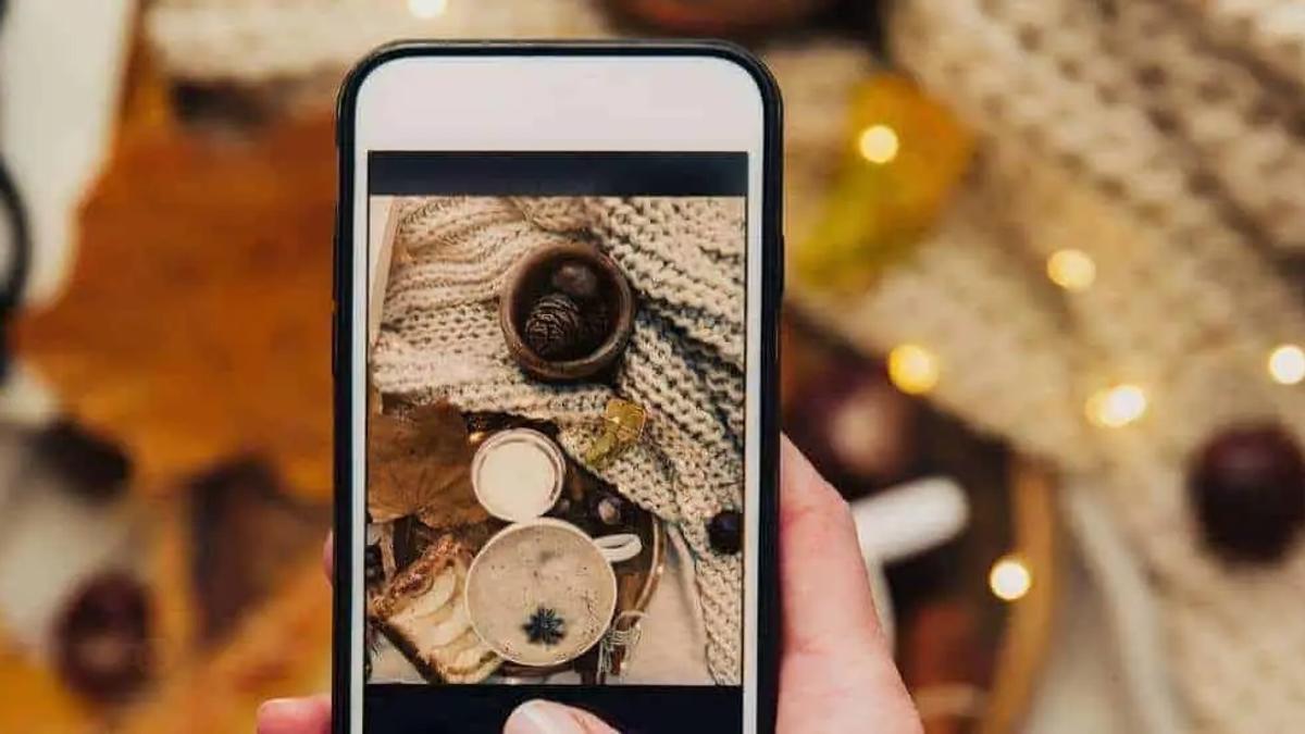 A hand holds a phone photographing a cozy autumn flat lay with coffee and pine cones