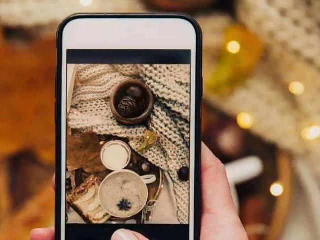 Una mano sostiene un telefono fotografiando una escena otonal con cafe y pinas
