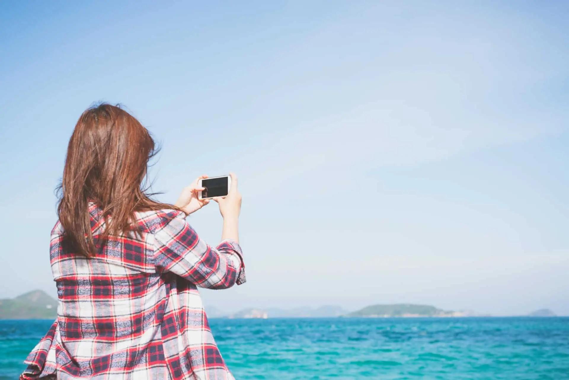 A woman in a plaid shirt takes a photo with her phone facing the ocean and distant hills