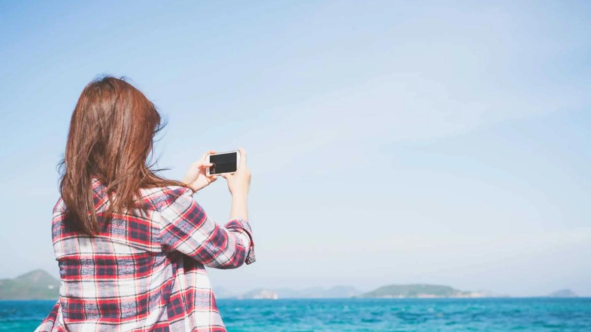 A woman in a plaid shirt takes a photo with her phone facing the ocean and distant hills