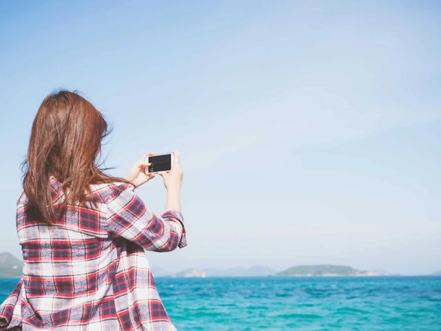 A woman in a plaid shirt takes a photo with her phone facing the ocean and distant hills