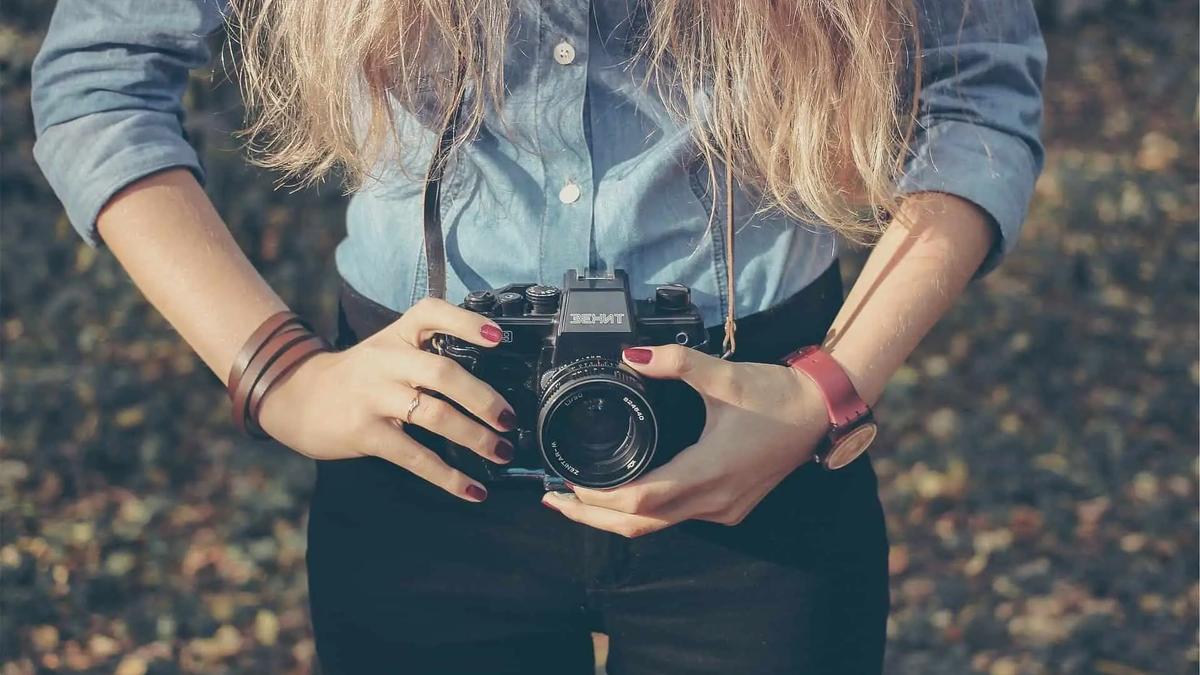 Person holding a black camera, wearing a blue shirt, black pants, brown bracelet, and red watch. Warm, soft lighting.