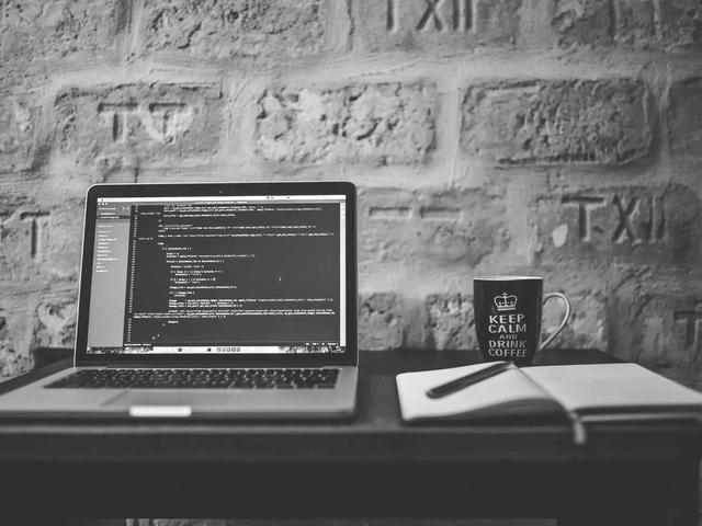 Black and white image of a laptop displaying code, a black mug with 'Keep Calm and Drink Coffee', and a notebook with a pen, on a dark table against a brick
